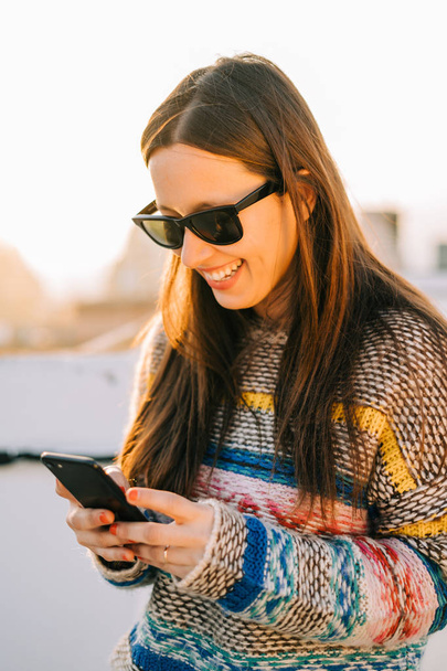 Young smiling woman with sunglasses using her smartphone on a terrace in New York City - Photo, image