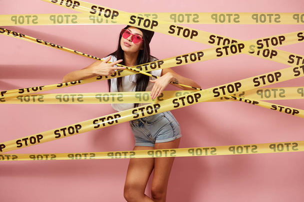 Dark-haired girl in pink sunglasses and cap dressed in jeans shorts and white top looking out from behind yellow scotch tapes with inscriptions of word "stop" on the background of pink wall - Zdjęcie, obraz