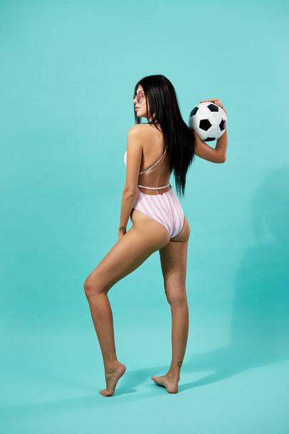 Slim dark-haired girl in sunglasses dressed in pink and white striped swimsuit  holds a soccer ball in her hands on the blue background - Foto, imagen