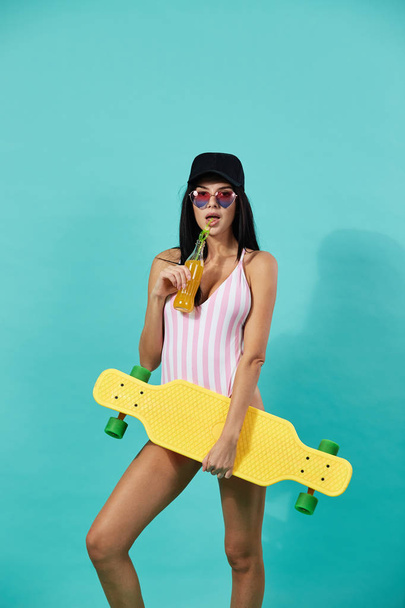 Slim dark-haired girl in sunglasses and cap dressed in pink and white striped swimsuit stands with yellow skateboard and bottle of juice in her hands on the blue background - Photo, Image