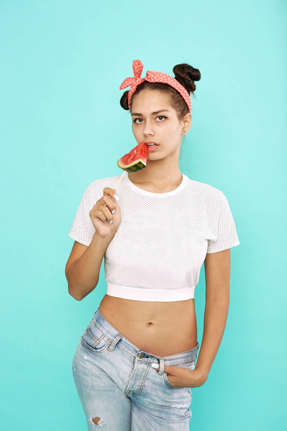 Young funny girl with pink bow on her head dressed in jeans and top is holding a slice of watermelon on a stick on the blue background in the studio - Photo, Image