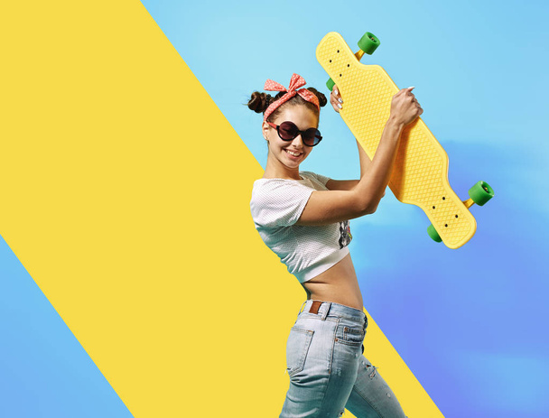 Young girl in sunglasses and pink bow on her head dressed in jeans and top stands with yellow skateboard on the back on the blue background in the studio - Foto, Imagem