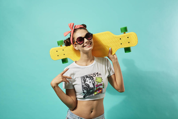 Happy young girl in sunglasses and pink bow on her head dressed in jeans and top stands with yellow skateboard on the blue background in the studio . . - 写真・画像
