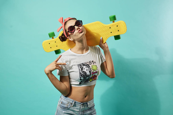 A napszemüveg és a fejét a rózsaszín orr boldog fiatal lány öltözött farmert és tetején áll a stúdióban kék alapon sárga gördeszka - Fotó, kép