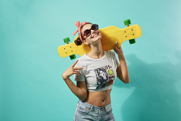 Happy young girl in sunglasses and pink bow on her head dressed in jeans and top stands with yellow skateboard on the blue background in the studio - Foto, Imagem