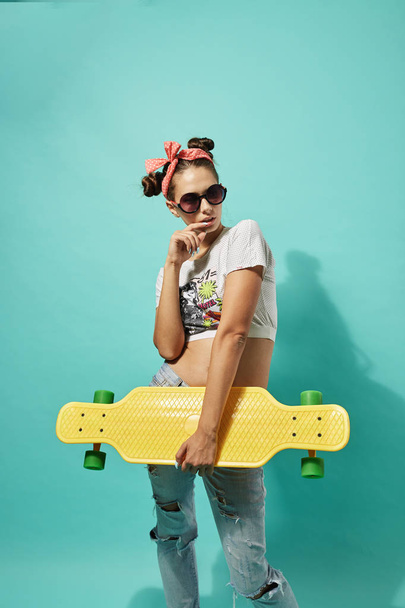 lustiges Teenie-Mädchen mit Sonnenbrille und rosa Schleife auf dem Kopf in Jeans und Top-Ständer mit gelbem Skateboard auf blauem Hintergrund im Studio - Foto, Bild