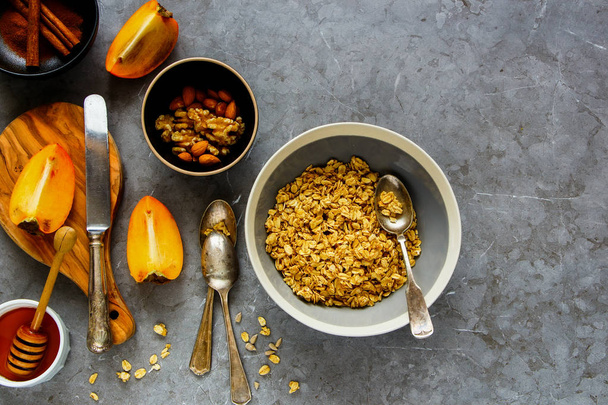 Flat-lay of healthy breakfast variety. Cinnamon granola bowl, fresh persimmon, honey and nuts over grey concrete table background, top view. Clean eating, dieting, vegetarian food - Image - Foto, immagini