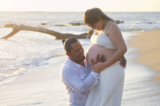 Ισπανόφωνος έγκυος γυναίκα με άντρα που ψάχνετε στην κοιλιά στο ηλιόλουστος υπόβαθρο - Φωτογραφία, εικόνα