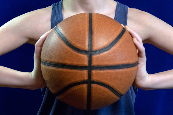 Θολή. Θέμα μπάσκετ. Μια έφηβη σε ένα αμάνικο μπλουζάκι κατέχει την μπάλα με τα δύο χέρια στο επίπεδο στήθος. Γκρο πλαν. Απομόνωση σε μπλε φόντο. - Φωτογραφία, εικόνα