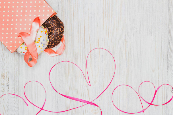 Gift bag with donuts inside and a red ribbon heart on a wooden table. Flat lay. Valentine's Day celebration concept. - Photo, Image