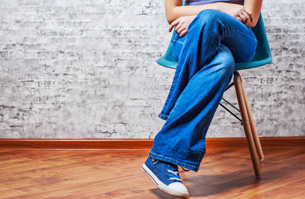 Woman fashion concept. Female legs in jeans and sneakers sits on a chair on gray wall background with copy space - 写真・画像