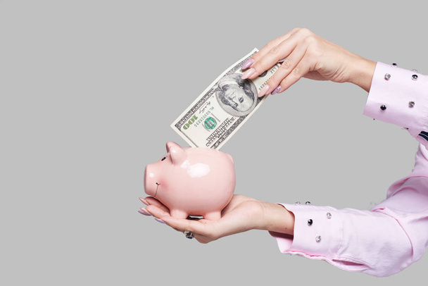 Cost of college education. Female student hand  puts cash in the piggy bank. The girl saves money for training in high school .Neutral grey background. Space for text - Photo, Image