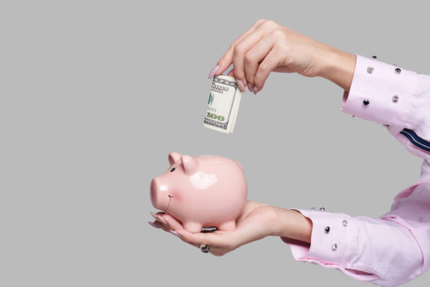 Cost of college education. Female student hand  puts cash in the piggy bank. The girl saves money for training in high school .Neutral grey background. Space for text - Photo, Image