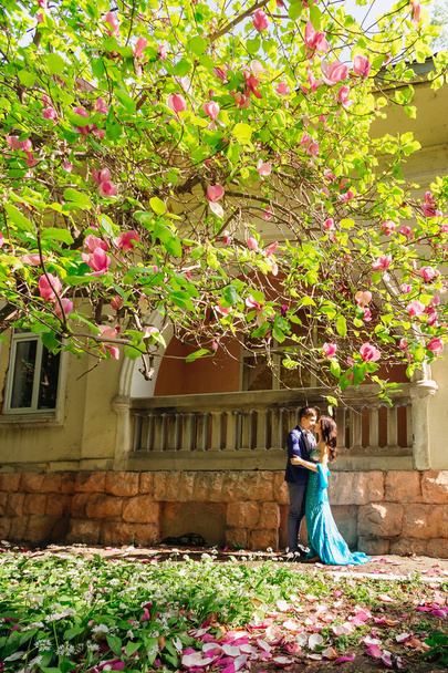 chico y chica de pie cara a cara cerca de un edificio con balcón. Ramas de magnolia con flores y pétalos de flores en el suelo
 - Foto, Imagen