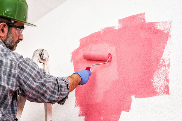 Peintre de maison caucasien travailleur sur une échelle en métal, avec le rouleau, il peint le mur avec la peinture colorée de rose. Industrie de la construction. Vue du bas
. - Photo, image