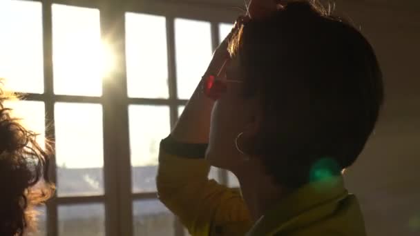 Μελαχρινή γυναίκα με γυαλιά ηλίου που ποζάρει πίσω στη φωτογραφική μηχανή μπροστά από το παράθυρο στο φως του ήλιου - Πλάνα, βίντεο