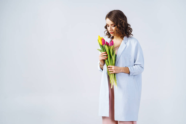 Fashion photo of a beautiful young woman with tulips in her hand .she dressed in a beautiful coat, and T-shirt with stripes.Spring concept. March 8. beautiful girl in stylish clothes - Фото, зображення