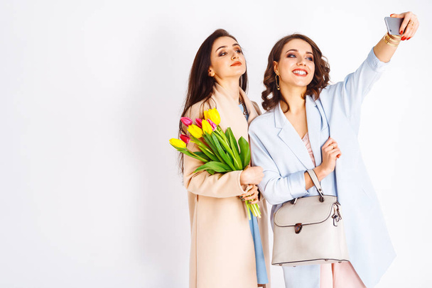 Fashion photo of two beautiful young women with tulips in the hand .they dressed in a beautiful coat, and T-shirt with stripes.Spring concept.March 8. beautiful girls in stylish clothes - Fotó, kép