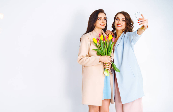 Fashion photo of two beautiful young women with tulips in the hand .they dressed in a beautiful coat, and T-shirt with stripes.Spring concept.March 8. beautiful girls in stylish clothes - Фото, зображення