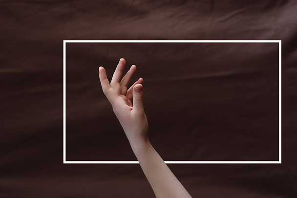 Female hand on the background of brown fabric with a drawn white frame. Minimal creative abstract concept. Flat lay. Copy space for text - Fotografie, Obrázek