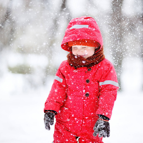 Portrait of little funny boy in red winter clothes having fun with snow during snowfall. Active outdoors leisure with children in winter. Kid with warm hat, hand gloves and scarf - Foto, imagen