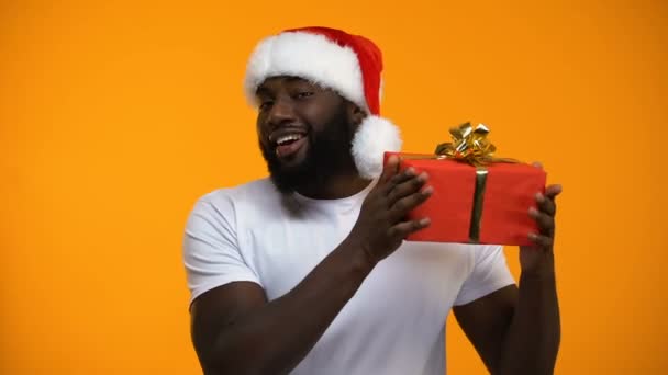 Afro-American man in Santa hat holding Christmas gift and winking, holiday - Metraje, vídeo