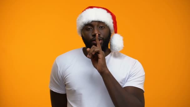 Black Santa showing silence sign and winking, holding gift, secret Xmas party - Кадри, відео