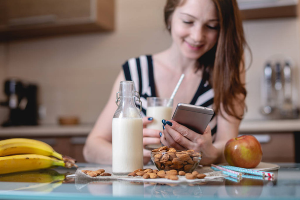 Женщина пьет натуральное миндальное молоко, держа телефон в руке на кухне. Бутылка и фрукты на столе. Вегетарианский продукт
 - Фото, изображение