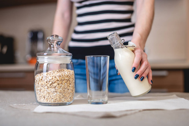 Uma mulher derrama leite de aveia orgânico de uma garrafa em um copo na cozinha. Dieta produto vegetariano saudável
 - Foto, Imagem