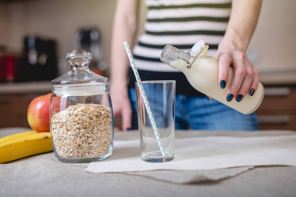 Una mujer vierte leche de avena orgánica de una botella en un vaso en la cocina. Dieta saludable producto vegetariano
 - Foto, Imagen