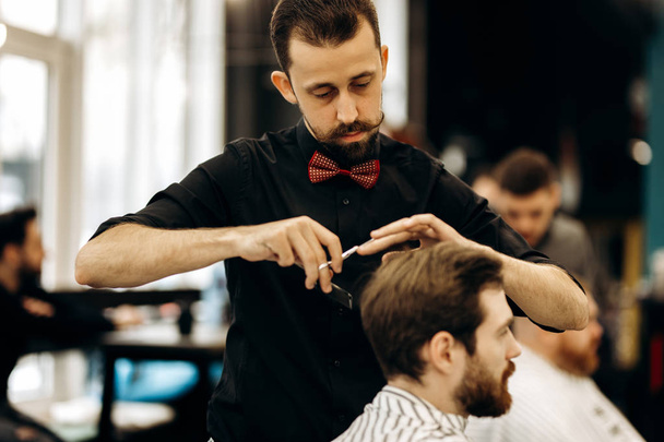  harismatic barber with mustache dressed in a black shirt with a red bow tie scissors the hair of a young man in a barbershop - Foto, Imagem