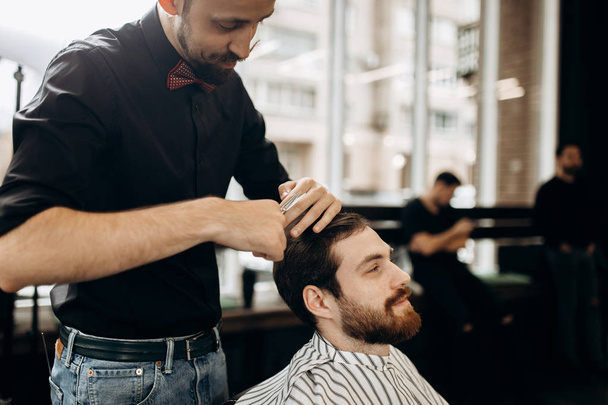 Mustachioed barber dressed in a black shirt with a red bow tie makes a stylish hairstyle to young man in a barbershop - Fotó, kép