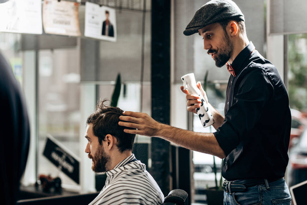 Mustachioed barber dressed in a black shirt, red bow tie and flat cap is doing the hair styling to the man sitting in the armchair in a barbershop - Фото, изображение