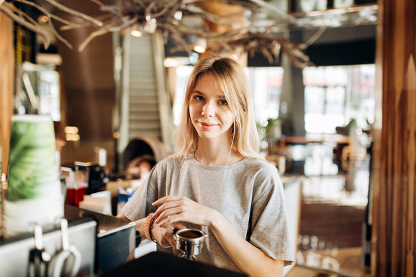 A pretty smiling slim girl,wearing casual clothes,holds some ground coffee and looks at the camera in a cozy coffee shop. - Fotografie, Obrázek