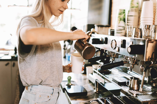 A youthful slim kind blonde girl,dressed in casual outfit,skillfully adds milk to coffee in a modern coffee shop. - 写真・画像