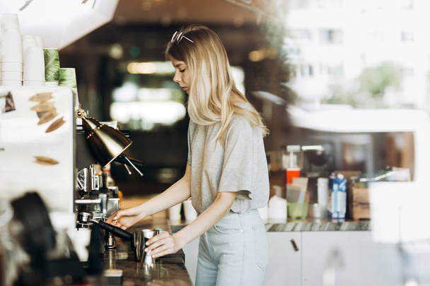 A young pretty thin blonde with long hair,dressed in casual outfit,is cooking coffee in a modern coffee shop. Process of making coffee is shown. - Фото, изображение