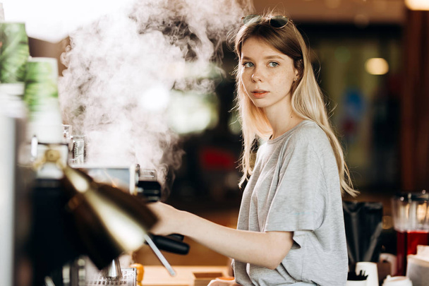 A youthful blonde slim gir,dressed in casual outfit,cleans the coffee machine with steam in a modern coffee shop. - Foto, Bild