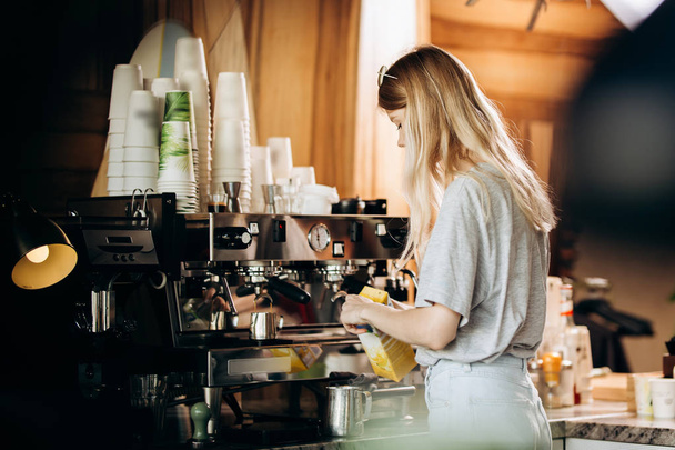 A good looking slim blonde with long hair,dressed in casual outfit,is cooking coffee in a modern coffee shop. Process of making coffee is shown. - Фото, изображение