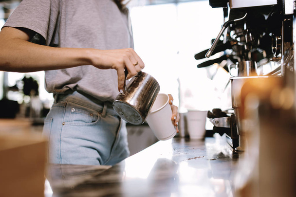 Una joven rubia delgada, vestida con ropa casual, se muestra añadiendo leche al café en una acogedora cafetería
. - Foto, Imagen