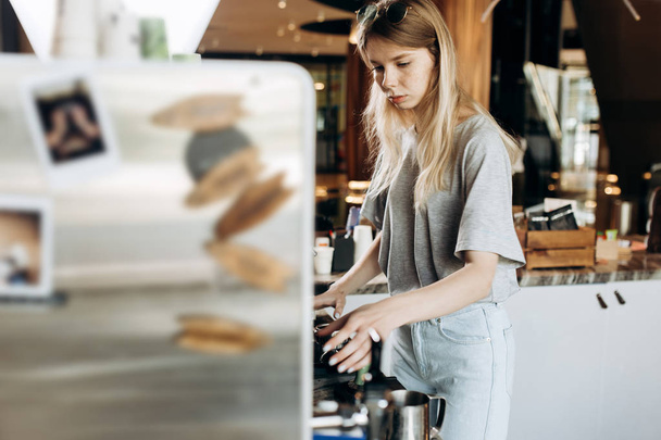 A young pretty thin blonde,dressed in casual outfit,is cooking coffee in a popular coffee shop. - Fotografie, Obrázek