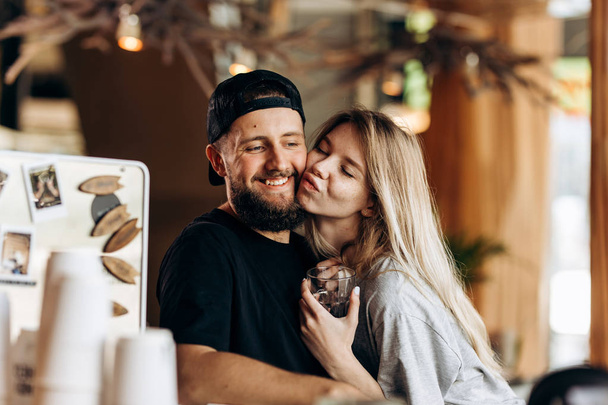 Dva mladí usmívající se lidi, blond dívka a muž s beard.dressed v neformální oblek, postavit vedle sebe a úsměv do kamery v útulné kavárně. - Fotografie, Obrázek