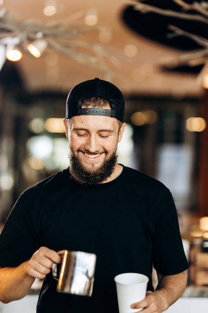 A handsome smiling young man with beard,dressed in casual style,adds milk to the coffee in a modern coffee shop. - Φωτογραφία, εικόνα