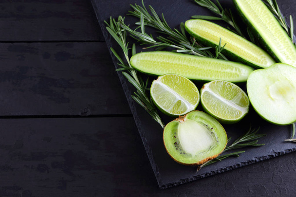 Green fruits, vegetables and rosemary on a black background with copy space. Creative figure of natural green products. Cucumbers, kiwi, lime and green apple on a slate board - Foto, immagini