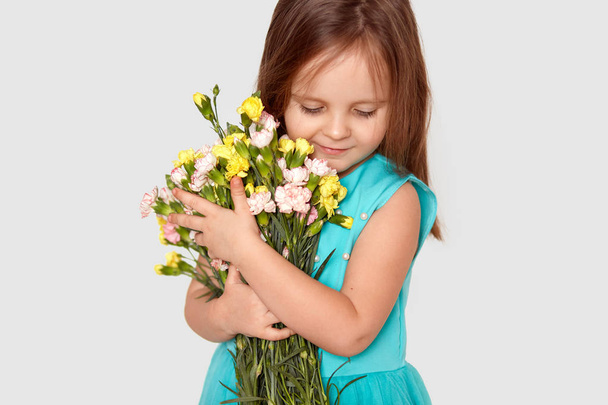 Cropped shot of pleased small Caucasian child with dark hair, focused down, carries beuatiful flowers, dressed in dress, recieves bouquet from father on 8 March, isolated over white background - Photo, image