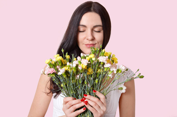Beautiful young lady with straight long hair, smells flowers with pleasant odour, closes eyes, wears white dress, has red manicure, isolated over rosy background. Woman and spring time concept - 写真・画像
