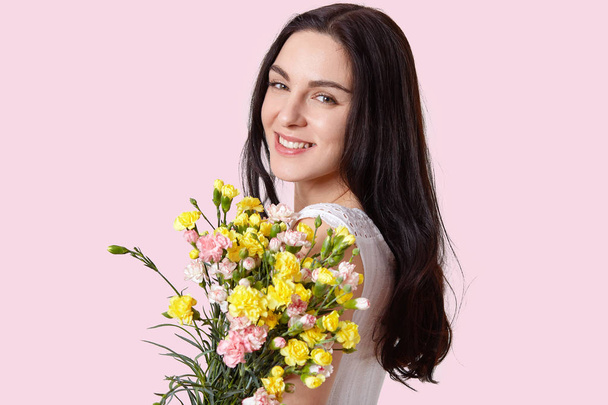 Headshot of pretty European young woman with tender smile, healthy skin, dark long hair, carries bouquet of spring flowers, looks at camera with happiness, isolated over light pink background - 写真・画像