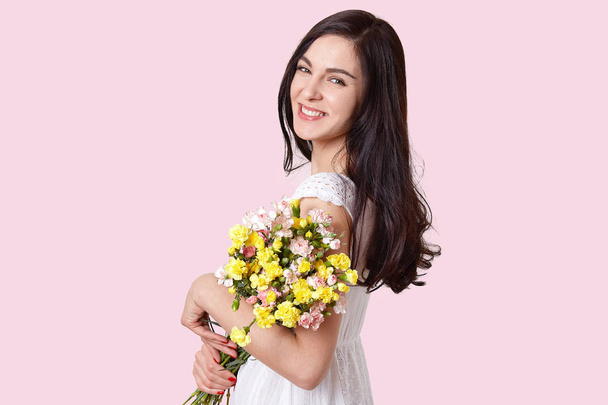 Photo of attractive brunette young woman with pleasant smile, dressed in white dress, holds bouquet of flowers, looks directly at camera, poses over light pink background, has minimal make up - Photo, image