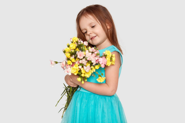 Sideways shot of attractive small child with long straight hair, dressed in blue stylish dress, looks positively at beautiful bouquet of flowers, isolated over white background. Children concept - Photo, image