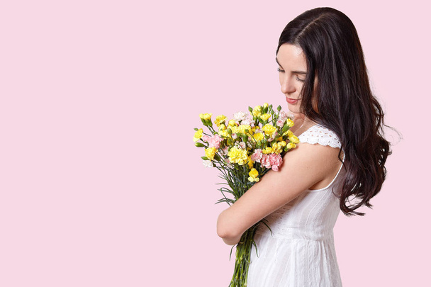 Studio shot of brunette woman looks at spring flowers, stands sideways, enjoys spring time, dressed in white dress, isolated over rosy background with empty space for your promotional content - Foto, imagen