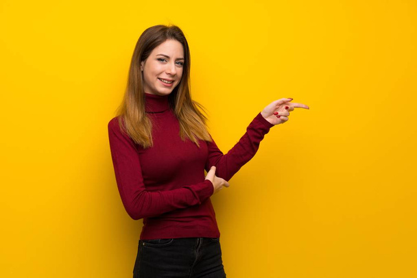 Nainen, jolla on poolokaula yli keltainen seinä osoittaa sormella sivulle
 - Valokuva, kuva